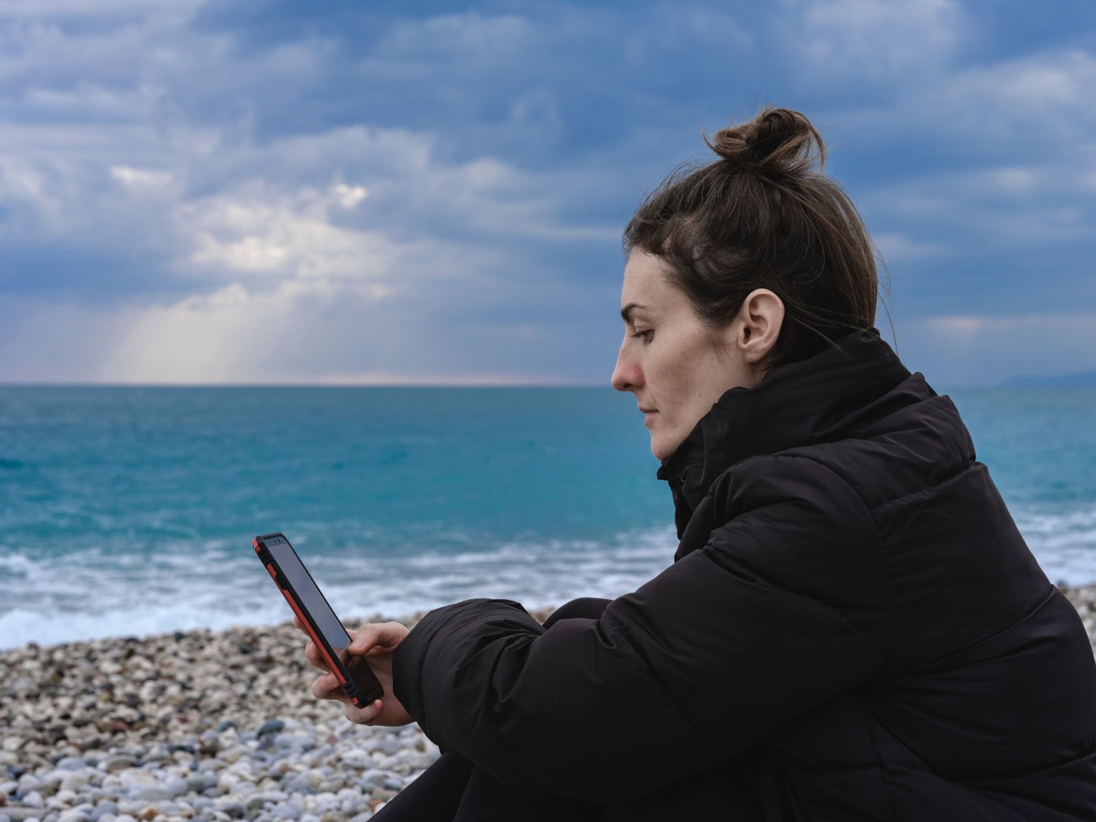 A woman on her phone in thought, sitting next to the ocean
